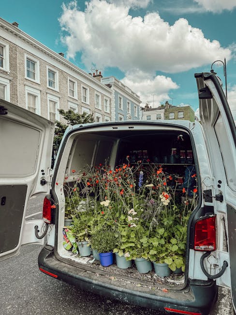 The image shows the open rear of a delivery van filled with a variety of fresh flowers and potted plants, including red poppies, white daisies, and green foliage, arranged in blue plastic pots and loo
