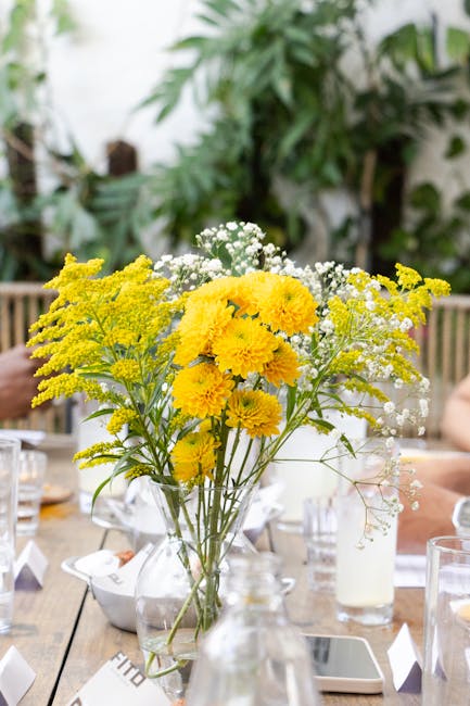 A floral arrangement featuring a bouquet of bright yellow chrysanthemums, delicate white baby's breath, and sprigs of yellow solidago, arranged in a clear glass vase. The bouquet is placed on a wooden