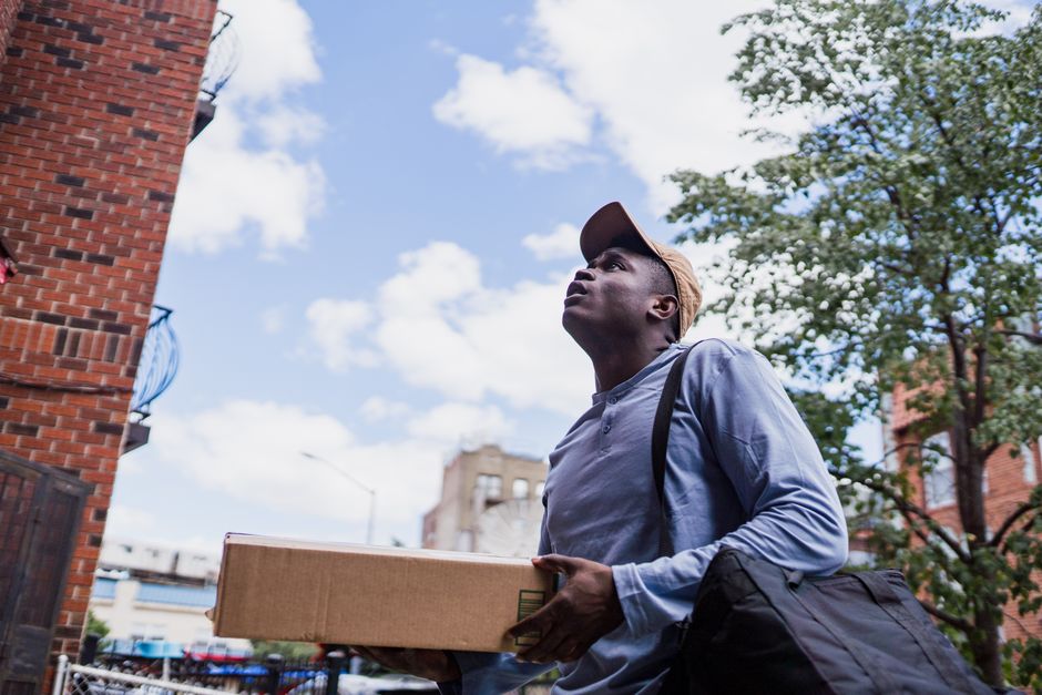 A young man outdoors in Kentish Town, holding a rectangular cardboard box, likely a floral gift or flower delivery package, with a neutral expression. He is wearing a light blue button-up shirt, a bro