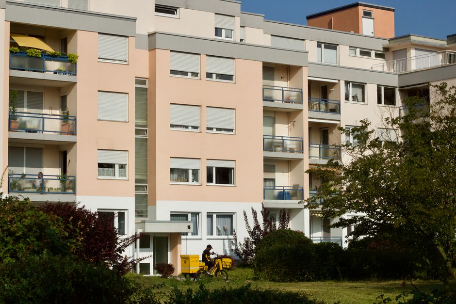 A modest flower delivery is being made outside a contemporary multi-story apartment building with a pastel pink and beige facade, featuring multiple balconies with glass railings. The delivery person,
