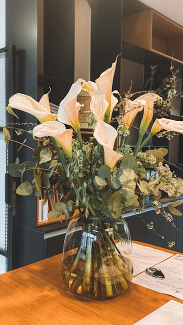 A tall, elegant floral arrangement featuring creamy white calla lilies with smooth, trumpet-shaped petals, complemented by sprigs of delicate white baby's breath and lush eucalyptus leaves. The flower
