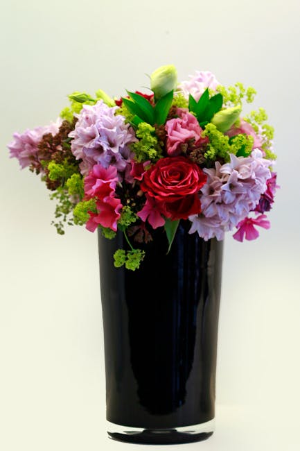 A vibrant floral arrangement in a tall, glossy black vase featuring an assortment of fresh flowers including a central red rose, pink and purple hydrangeas, white tulips, and small green foliage accen