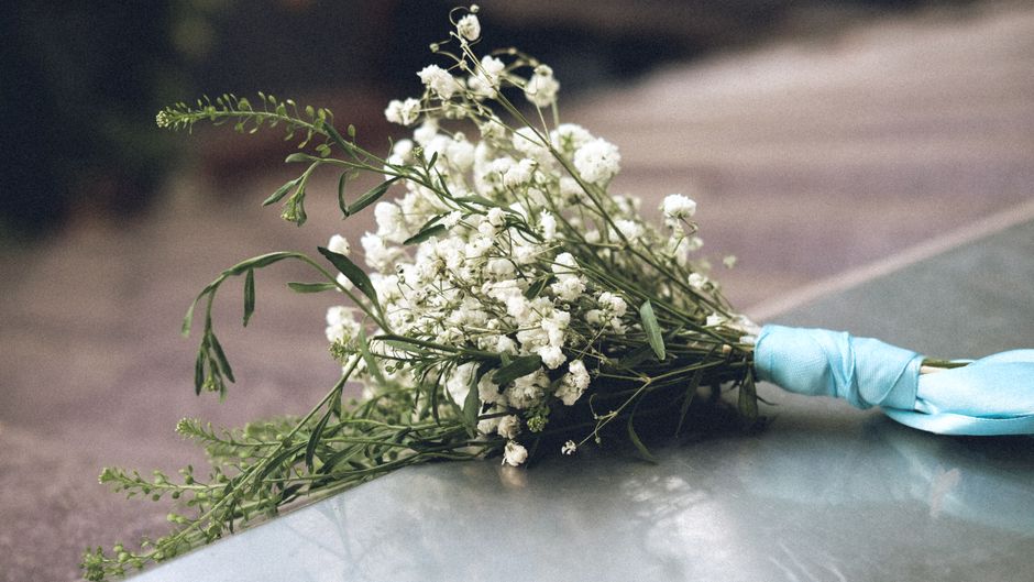 A delicate bouquet of white baby's breath flowers with small, round clusters and slender green stems, wrapped in light blue satin ribbon. The bouquet is resting on a smooth, gray surface with soft nat