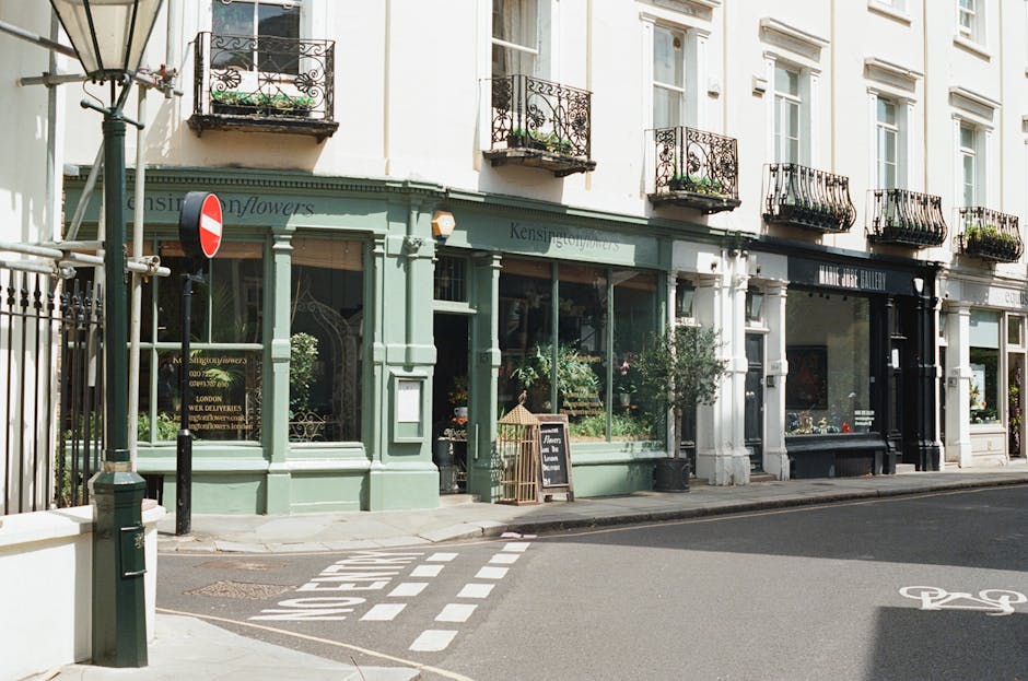 A row of shopfronts on a city street featuring a florist shop with a mint green exterior, large glass display windows filled with potted plants and floral arrangements, and a blackboard sign outside a