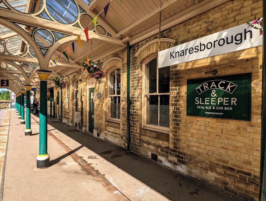 A detailed image of Knaresborough station platform showing vintage brick station building with arched windows and a green sign reading 'Track & Sleeper Real Ale & Gin Bar'. The platform is covered by