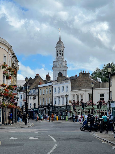 A bustling street scene in Kentish Town during daytime, featuring historic terraced buildings adorned with hanging flower baskets and shopfronts. The prominent white clock tower with a gold weather va