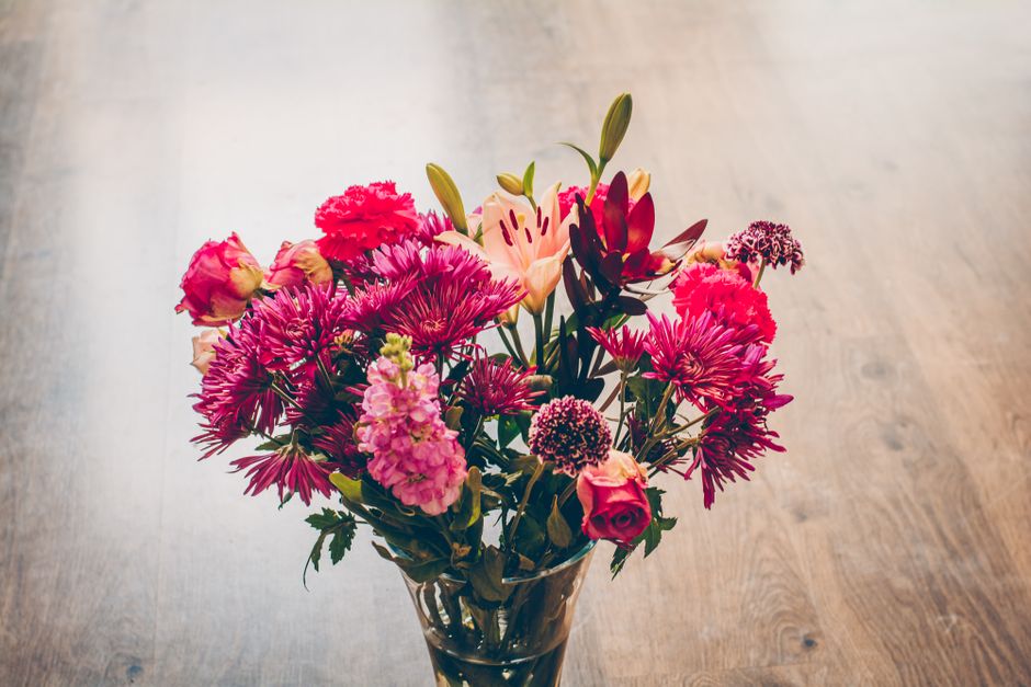 A vibrant bouquet of fresh flowers arranged in a clear glass vase, featuring pink and red roses, purple chrysanthemums, peach lilies, and small purple statice, with green foliage. The bouquet is place