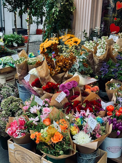 A vibrant display of fresh flowers arranged in bouquets at a flower shop window. The arrangement includes sunflowers, pink and purple lilies, red carnations, orange roses, and white chrysanthemums, al