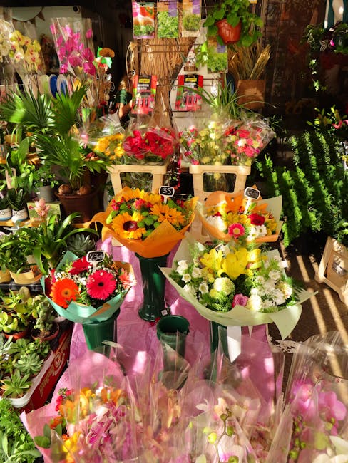 A vibrant display of fresh flower bouquets arranged on a pink cloth-covered table inside a flower shop. The bouquets include bright pink gerbera daisies, yellow and orange sunflowers, white roses, and