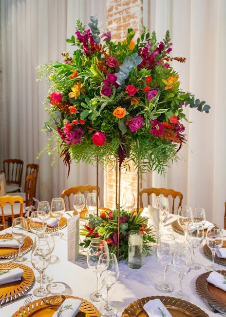 A large, vibrant floral arrangement featuring a mix of pink, purple, yellow, orange, and green flowers, including roses, delphiniums, and greenery, arranged in a tall glass vase on a table set for a f